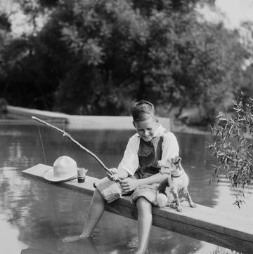 boy fishing with homemade pole, feet dangling in water, dog sitting by his side photo by h armstrong robertsretrofilegetty images