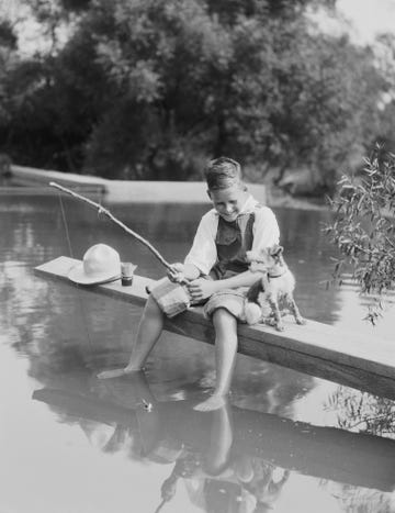 boy fishing with homemade pole, feet dangling in water, dog sitting by his side photo by h armstrong robertsretrofilegetty images