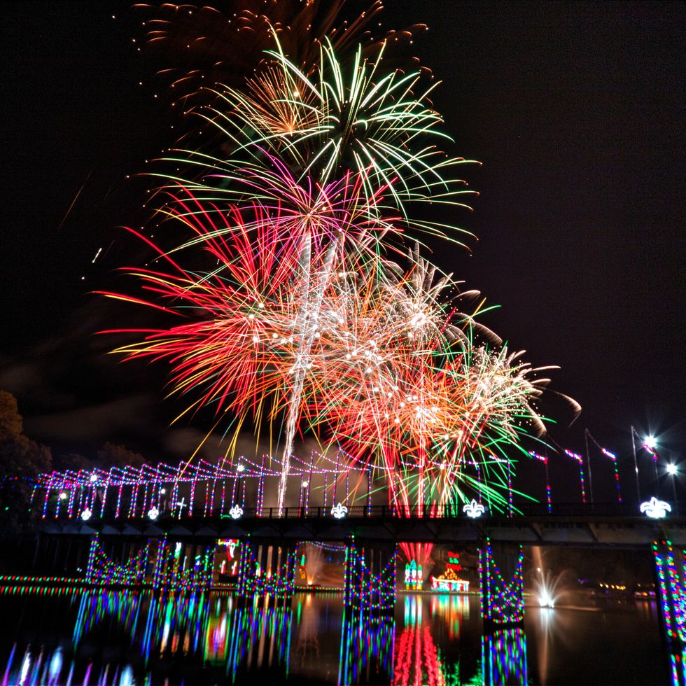 fireworks bursting over river with bridge lighted below it in christmas lights
