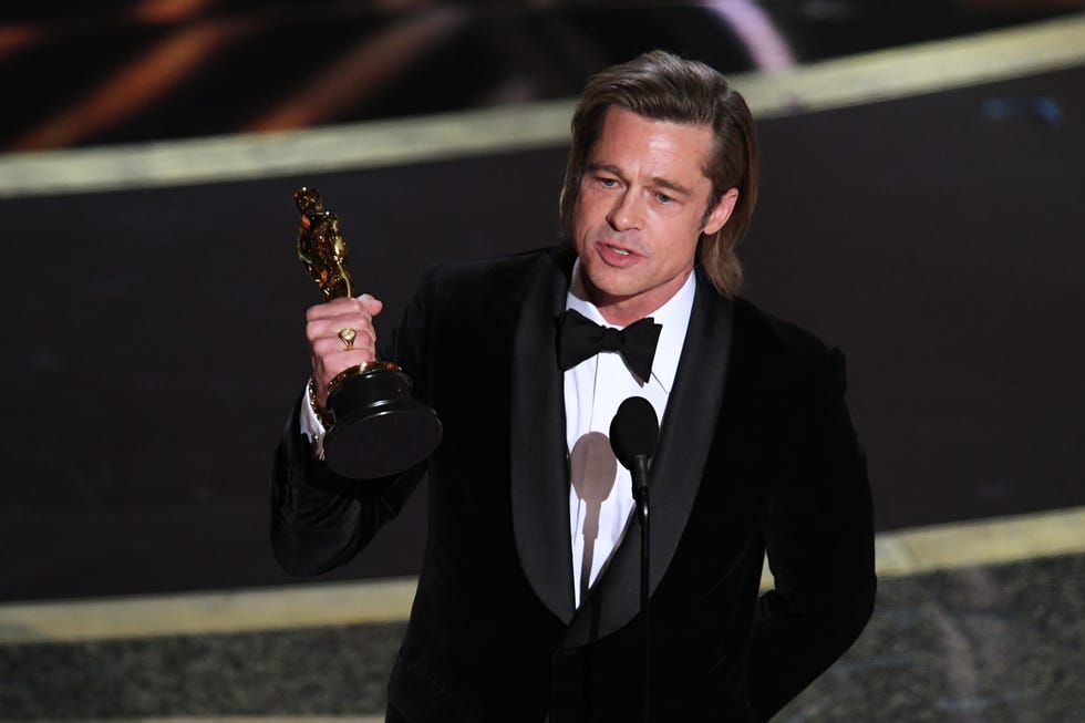 brad pitt holds oscar in hands as he speaks into a microphone, he wears a black suit jacket, bow tie and white collared shirt