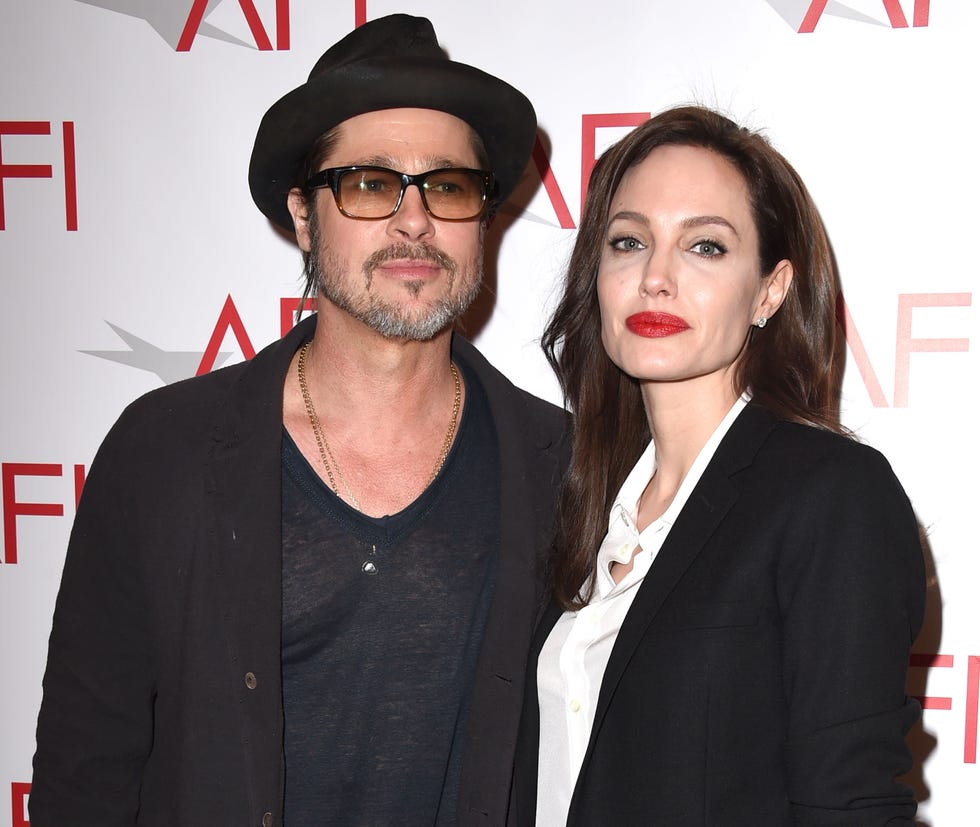 brad pitt and angelina jolie stand together and pose for photos, he wears a hat, sunglasses, a sweater and a tshirt, she wears a blazer and a white collared shirt