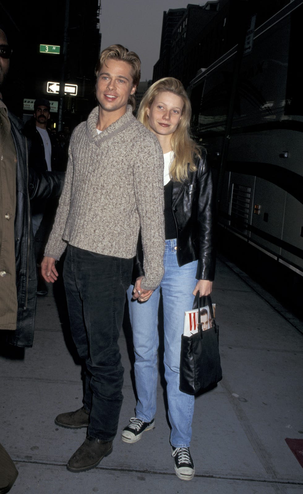 brad pitt and gwyneth paltrow﻿ hold hands while standing on a city street and next to a parked bus, he wears a chunky sweater and dark jeans, she wears a black jacket, sweater, white shirt and jeans