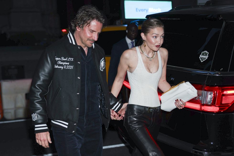 a man in a black letterman style jacket and a woman in a white tank top walk together outside at night, cars appear in the background