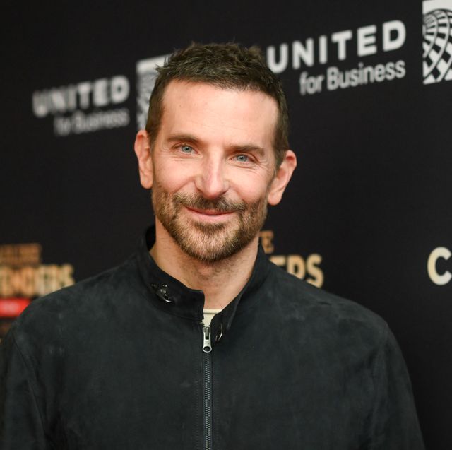 bradley cooper smiles at the camera and wears a black zip up jacket