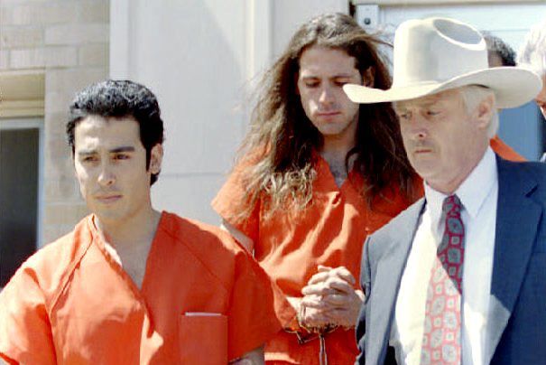a man wearing a blue suit and cowboy hat escorts two inmates in orange jumpsuits down a flight of stairs outside