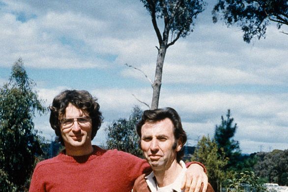 david koresh, wearing a red shirt and olive pants, smiles and puts his arm around the shoulders of clive doyle, wearing a brown shirt and blue jeans, as they stand in a grassy area in front of a road