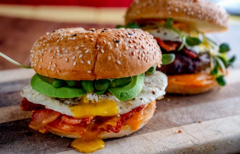 breakfast bagel with egg, greens, avocado, bacon, sausage and cheese on a rustic cutting board