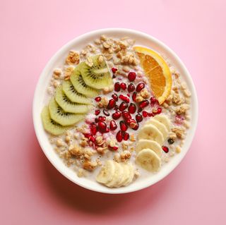 breakfast cereal meal granola with milk, pomegranate, kiwi and orange in bowl over pink background