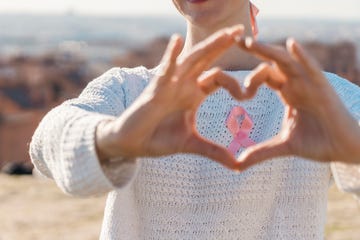 woman seen from chin to stomach holding out hands to make a heart shape to frame a pink ribbon on her white sweater