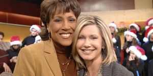 robin roberts and olivia newton john smile at the camera and embrace as they stand in front of several people wearing santa hats, roberts wears a tan suit jacket and brown turtleneck with jewelry, newton john wears a gray sweater and jewelry