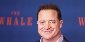 brendan fraser smiles at the camera, he wears a gray suit jacket and purple collared shirt that is unbuttoned, behind him is a blurred out blue background