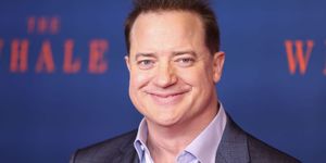 brendan fraser smiles at the camera, he wears a gray suit jacket and purple collared shirt that is unbuttoned, behind him is a blurred out blue background