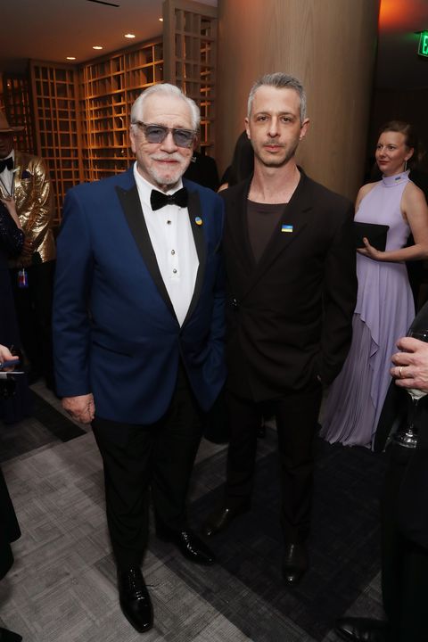 brian cox, wearing a blue tuxedo and sunglasses, stands next to jeremy strong, wearing a black suit jacket and black t shirt