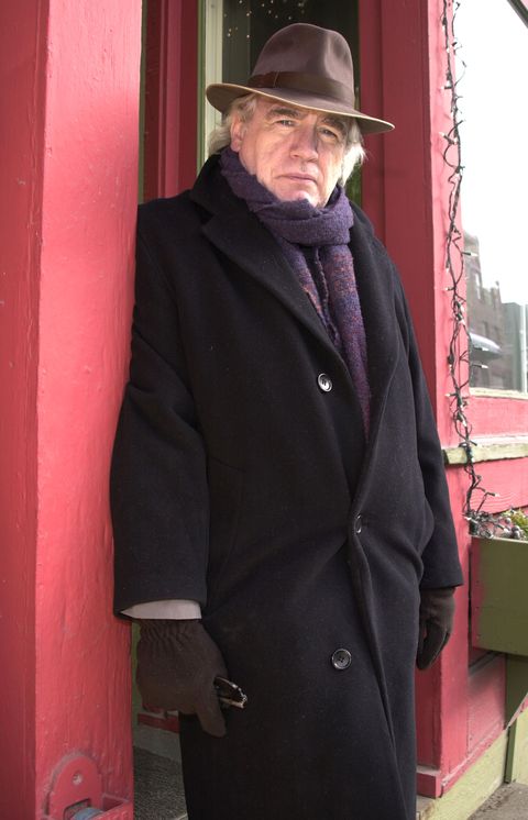 brian cox wearing a black coat, purple scarf, and brown hat, unsmiling and looking into the camera