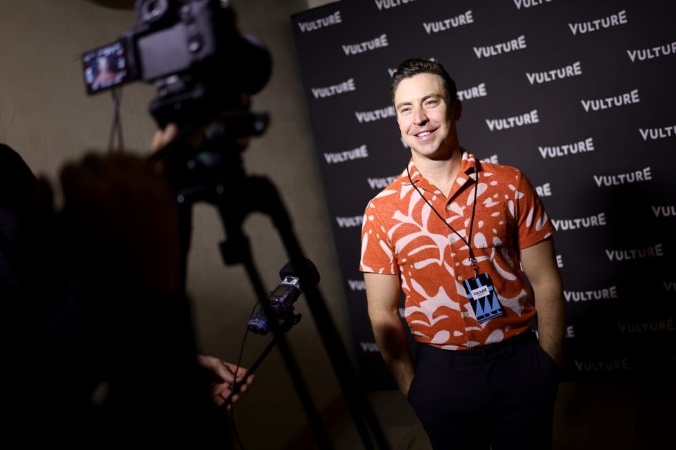 brian jordan alvarez smiles toward a camera to the left, he stands in front of a black background and wears an orange patterned button up short sleeve shirt, pants and a lanyard with an id