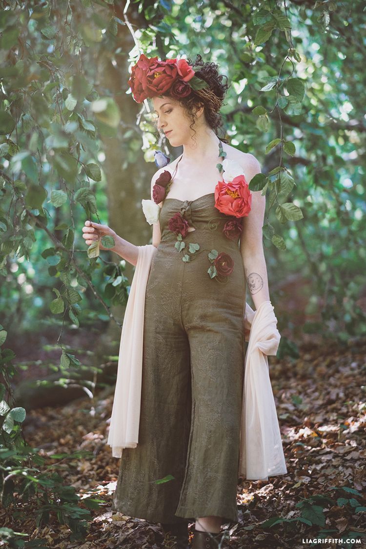 woman dressed as mother earth in a forest wearing a flower crown and flowers and ivy on her muted green jumpsuit she also has a gauzy cream wrap