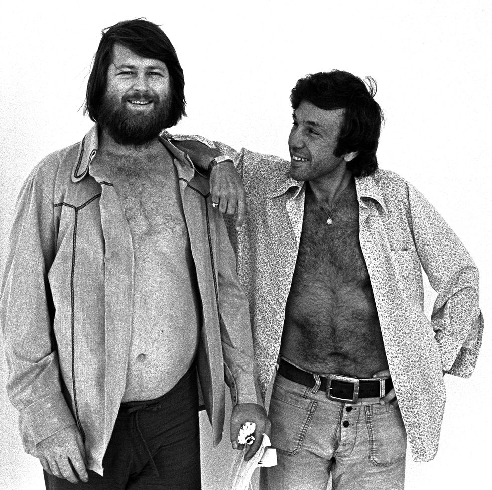 two men smile while standing together and wearing unbuttoned collared shirts that show their chests