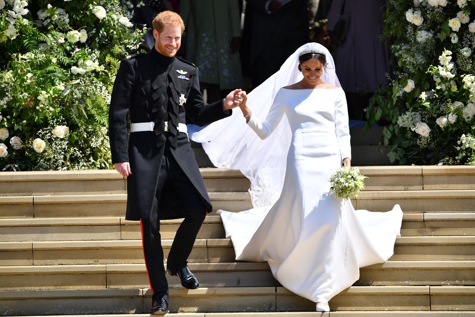 prince harry and meghan markle hold hands as they walk down stone steps, he wears a black military uniform, she wears a white wedding dress and veil