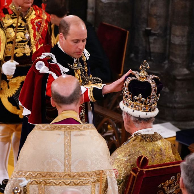 prince william stands before king charles who is seated and wearing a large crown, william touches the crown and looks down, both men wears robes as other men stand and watch