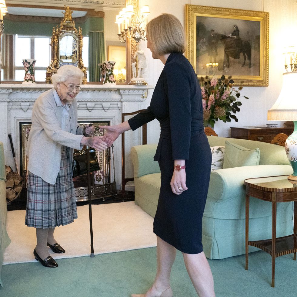 queen elizabeth ii shakes hands with liz truss as both women stand in a living room, elizabeth wears a gray cardigan, blue shirt, and plaid skirt, truss wears an all black skirt suit, the room has green carpet, two green couches and a fireplace with several decorations
