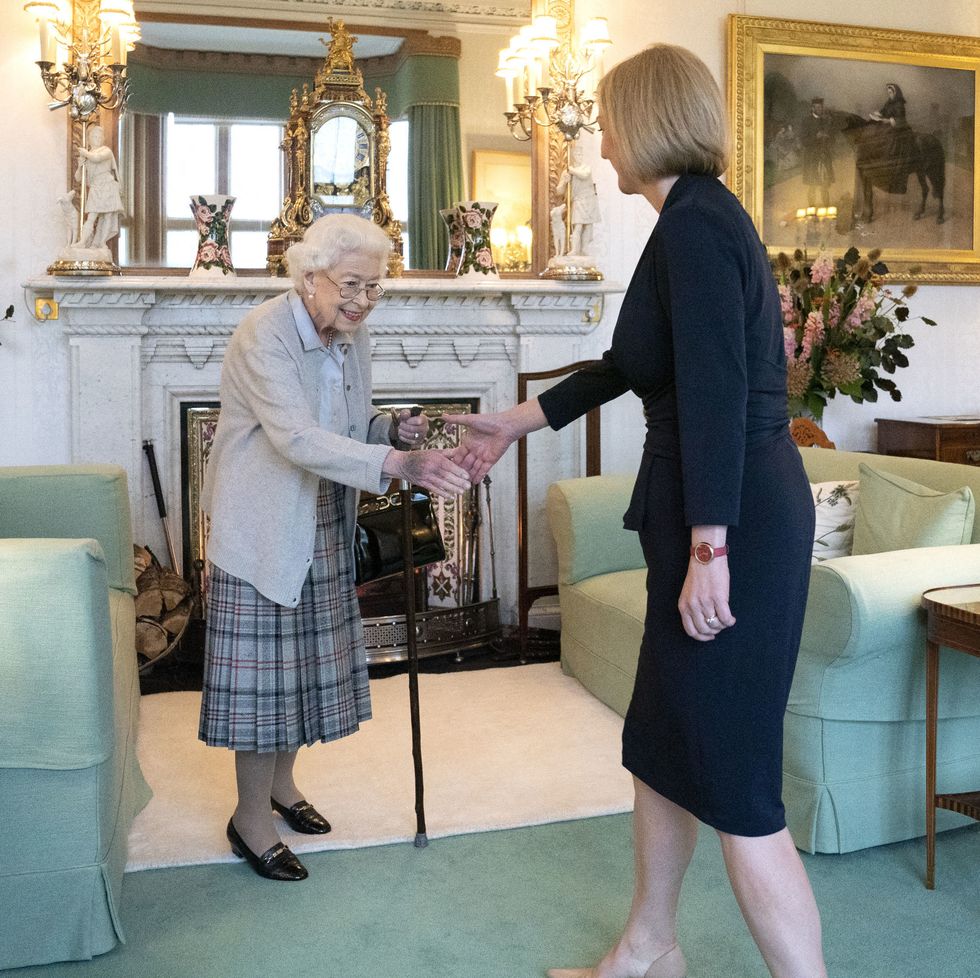 queen elizabeth ii shakes hands with liz truss as both women stand in a living room, elizabeth wears a gray cardigan, blue shirt, and plaid skirt, truss wears an all black skirt suit, the room has green carpet, two green couches and a fireplace with several decorations