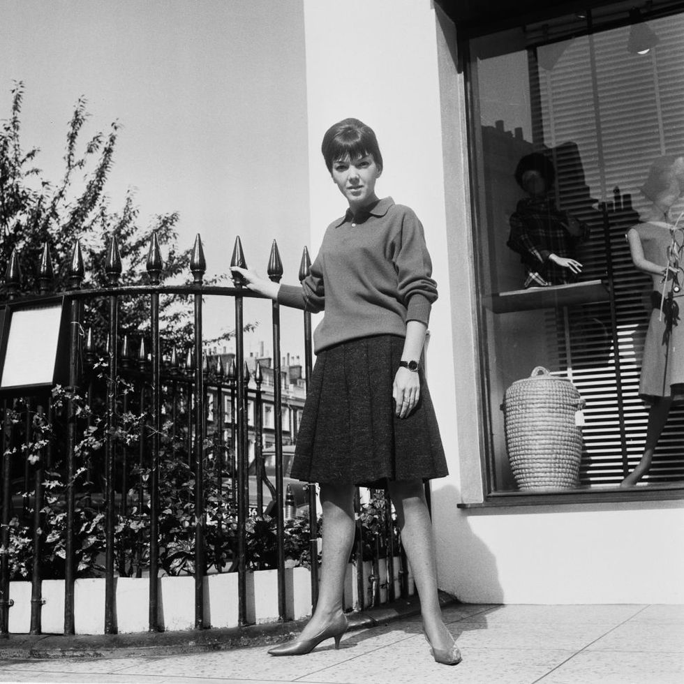 mary quant stands outside of her boutique bazaar, she is wearing a collared sweater and a knee length pleated skirt with short heels and a watch, her right hand holds a metal fence on her right and behind her is part of the storefront of her boutique