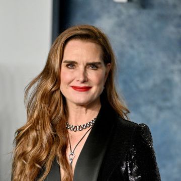 brooke shields smiles at the camera, she is wearing a black sequin suit jacket with large lapels and two necklaces, her wavy hair is styled down mostly over her right shoulder
