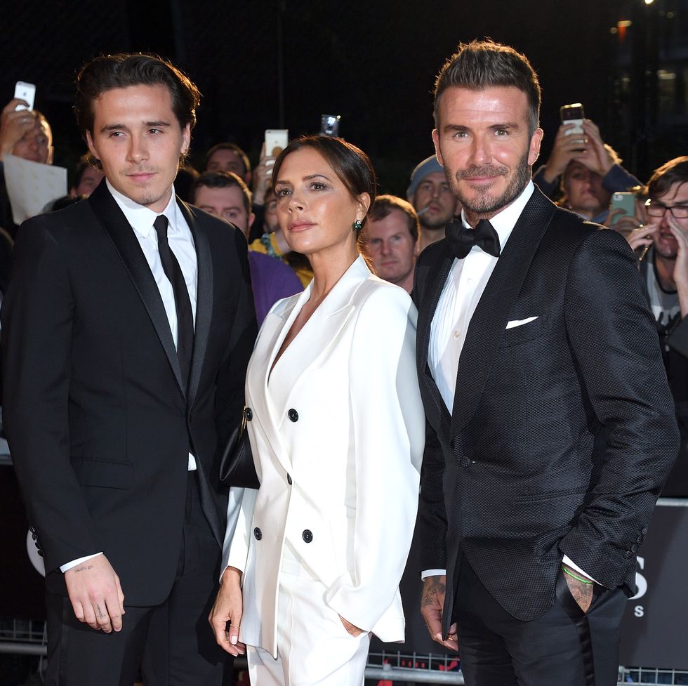 brooklyn beckham, victoria beckham, and david beckham stand for photos in front of a crowd of people, all three wear suits, the men in dark black and victoria in all white