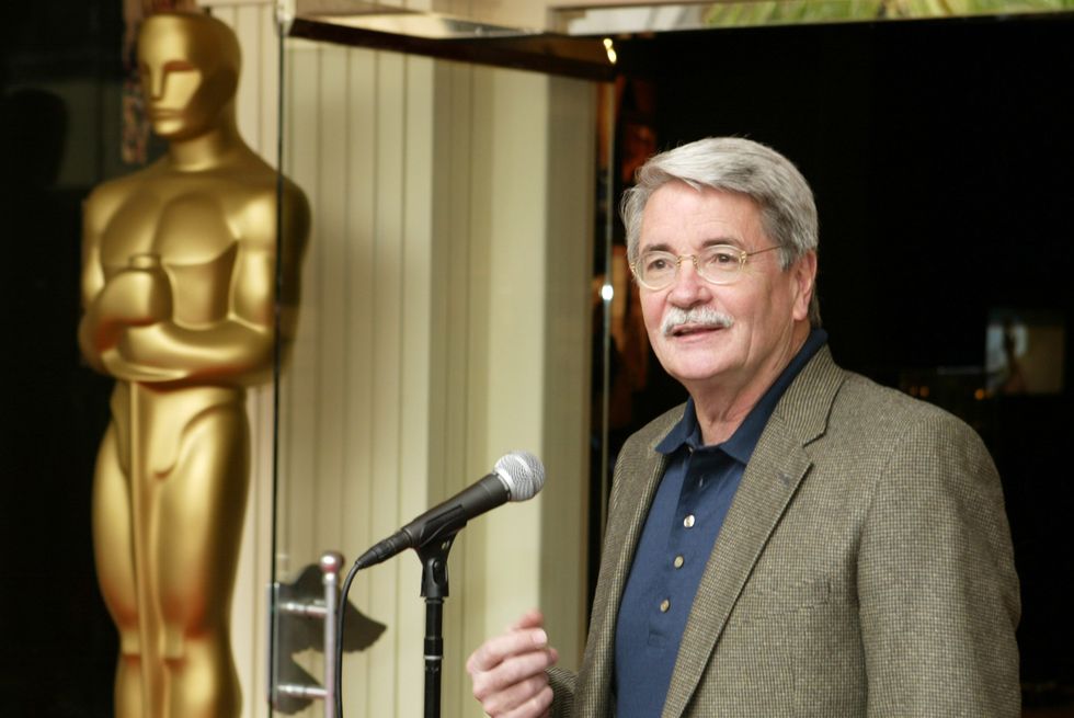 bruce davis, wearing a tan suit coat, blue shirt and glasses, stands in front of a microphone and speaks, with a large oscar statute replica behind him