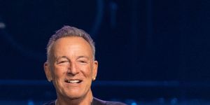 bruce springsteen smiles and stands while holding an electric guitar, he wears a navy t shirt