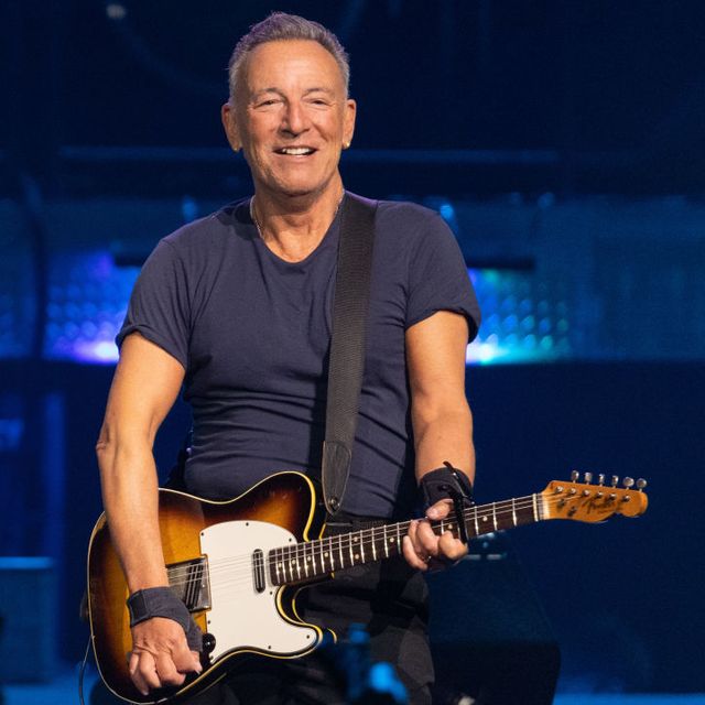 bruce springsteen smiles and stands while holding an electric guitar, he wears a navy t shirt