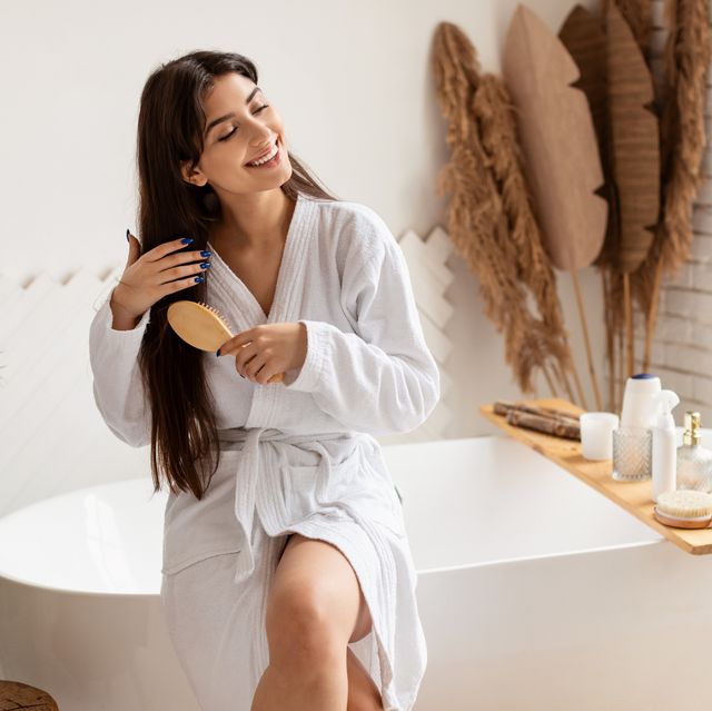brunette female brushing hair with hairbrush sitting on bathtub indoor