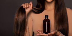 brunette girl with long straight and shiny hair　
beauty skin woman holding her strong and healthy hair and oil or shampoo bottle over grey background cosmetic hair beauty salon concept