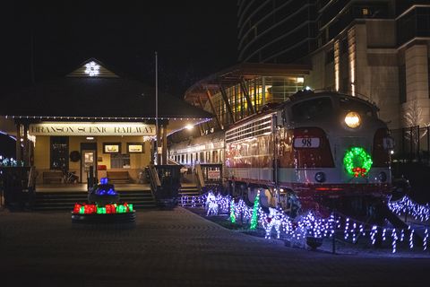 old fashioned red and white train with a lighted wreath on its front sitting in a station at night with blue lights around it the depot next to it has a sign that reads branson scenic railway