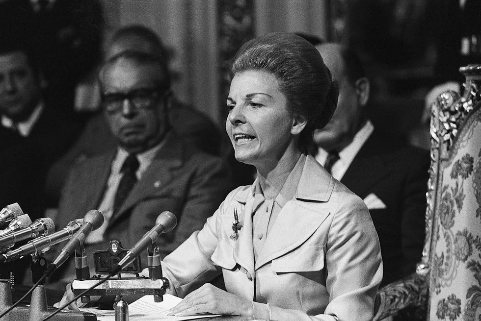isabel peron speaks into several microphones while seated at a table, she holds a stack of papers in front of her and wears a jacket and collared blouse