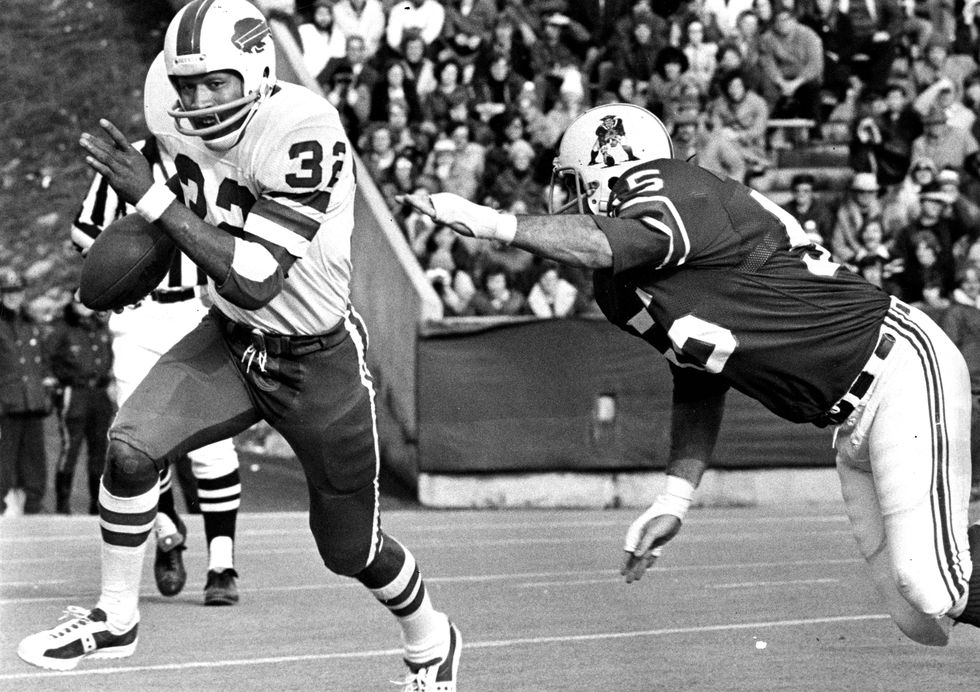 oj simpson holding a football in full uniform and running away from a defender during a game