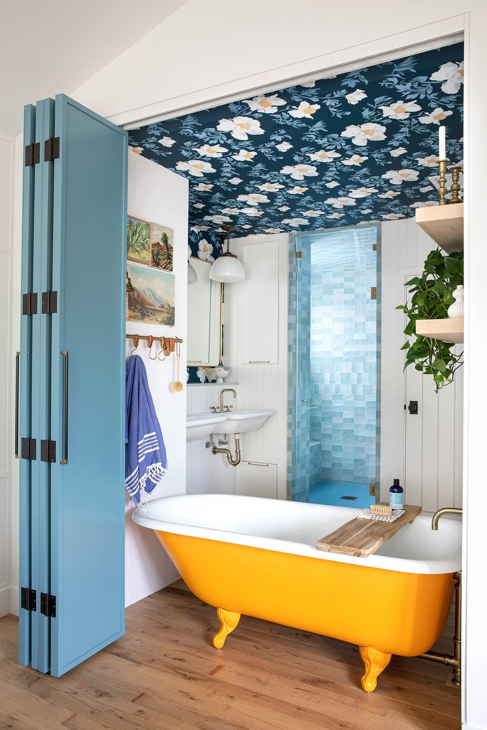 baño de estilo retro con bañera con patas amarilla, ducha, papel pintado floral en el techo y puertas plegables azules