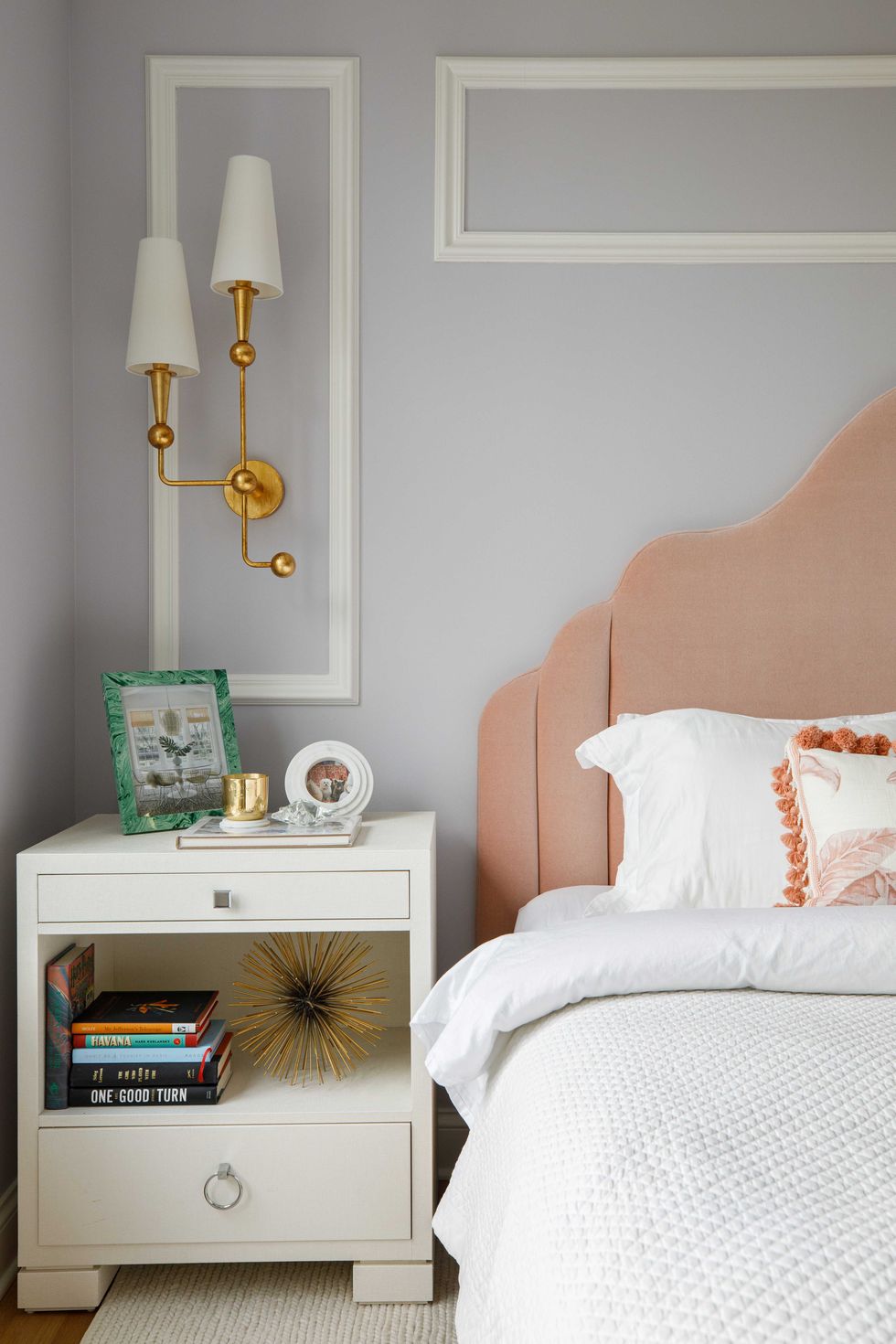bedroom, bed with white linon, white bed side table, books, pink headboard, white and gold sconces, white crown moulding on light grey blue painted walls