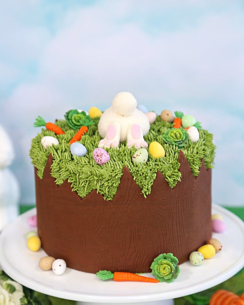 a cake decorated to look like a tree stump with piped grass and moss on the top with candy eggs and a bunnys fluffy tale and feet sticking up in the middle