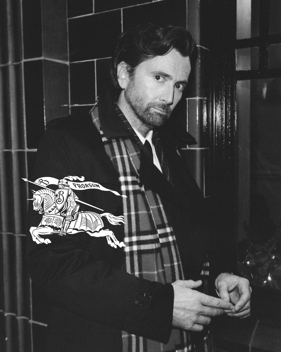 david tennant stands against a tiled wall wearing a burberry black coat and a plaid scarf