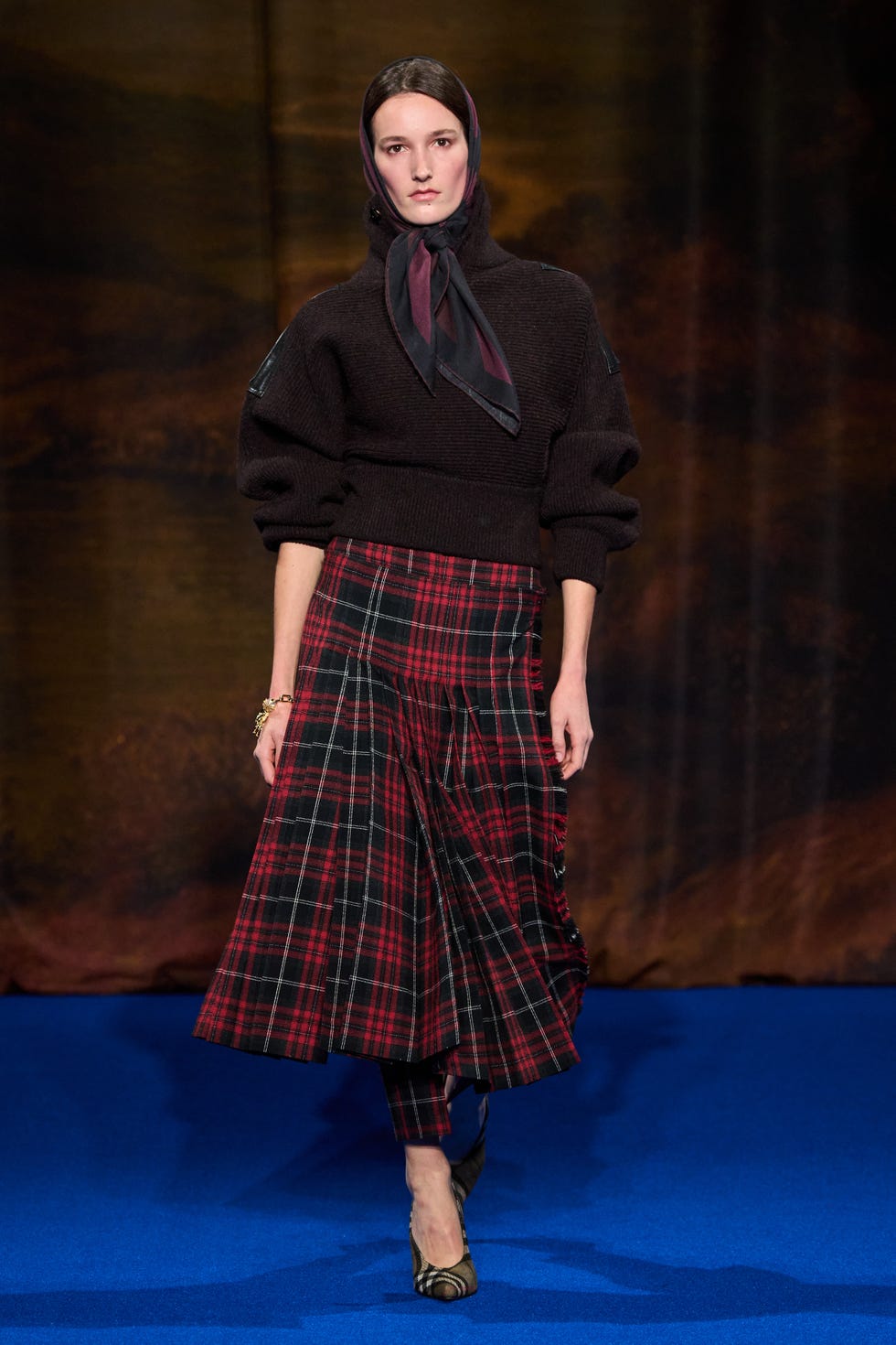 Female model showcasing a stylish outfit featuring a sweater and plaid skirt on a runway