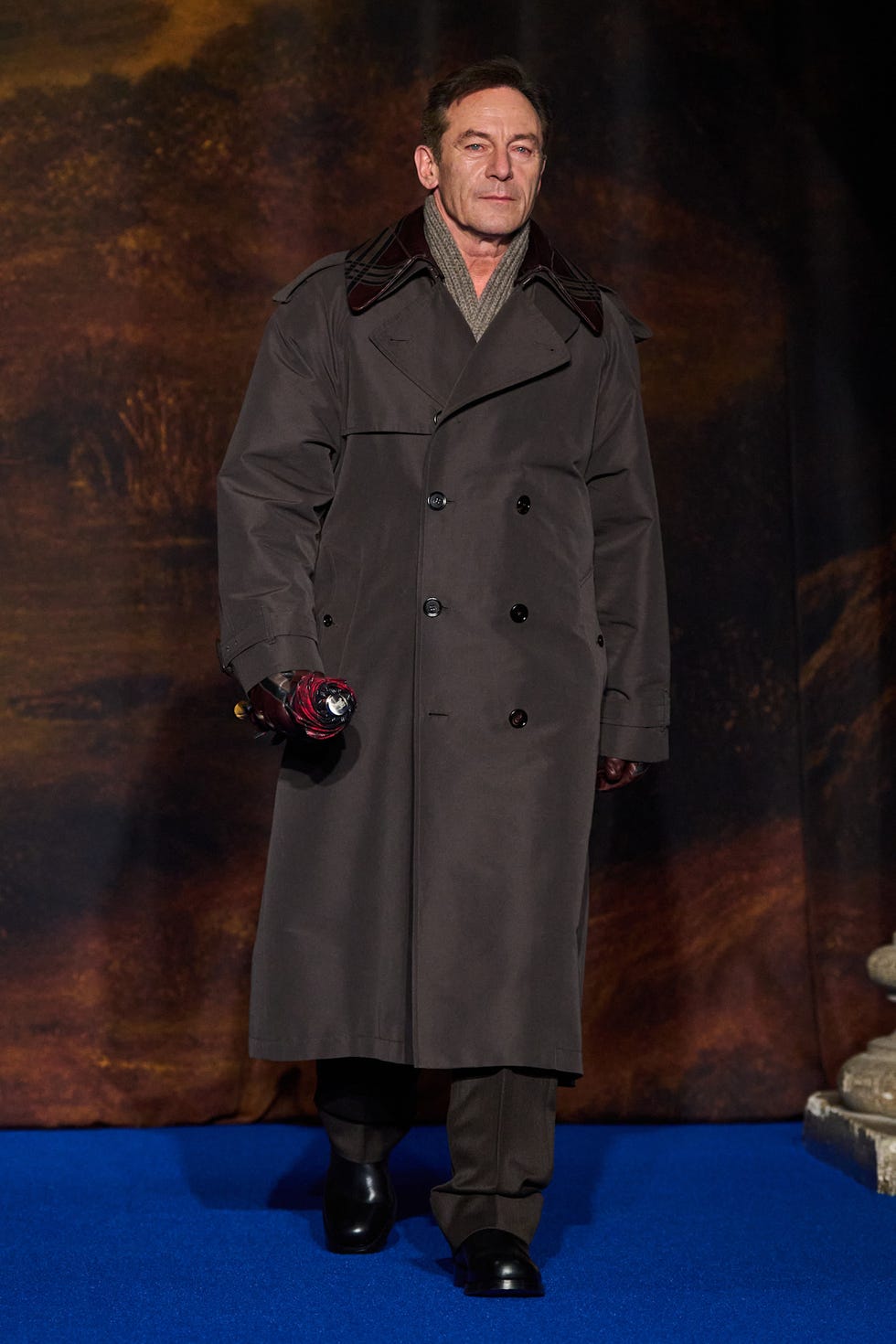a person dressed in a long coat holding an umbrella walking on a blue carpet
