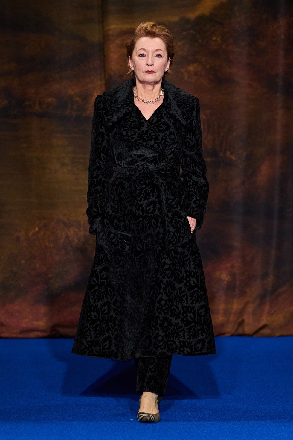 a person wearing a long dark coat with a floral pattern and matching trousers on a runway