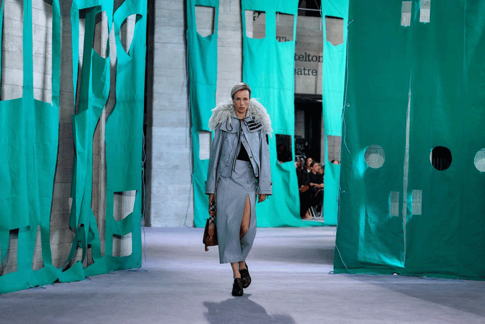 a model in a beige trench coat walking the runway in front of a green cloth art installation