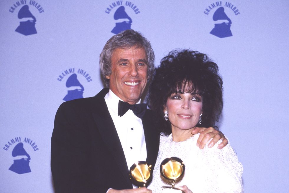 burt bacharach and carole bayer sager each hold a grammy award as they have one arm around each other and smile at a camera on their left, they are standing in front of a purple grammy awards backdrop, bacharach wears a black tuxedo and sager wears a white dress, large dangling earrings, and large bracelets