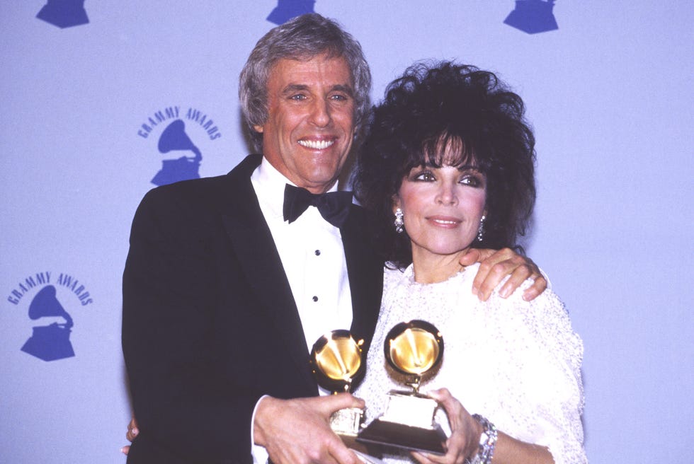 burt bacharach and carole bayer sager each hold a grammy award as they have one arm around each other and smile at a camera on their left, they are standing in front of a purple grammy awards backdrop, bacharach wears a black tuxedo and sager wears a white dress, large dangling earrings, and large bracelets