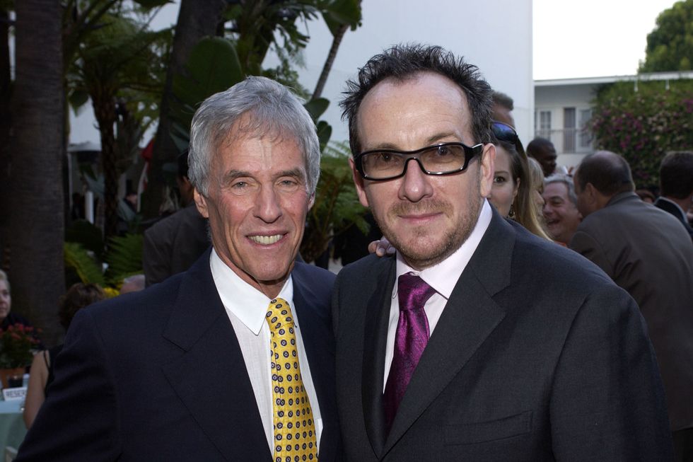 burt bacharach and elvis costello pose for a photo together, each with an around around the other, bacharach is wearing a navy suit coat, white collared shirt, and yellow and blue tie, while costello wears a black suit coat, white collared shirt, a purple tie, and tinted glasses with black frames, both men smile at the camera