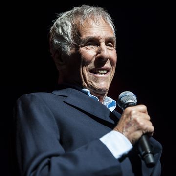 burt bacharach smiling and standing on a stage, holding a microphone and wearing a suit jacket