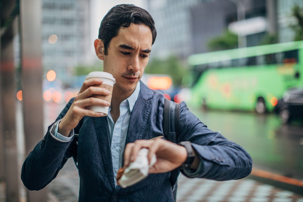 hombre comiendo en la calle y mirando el reloj con la mano que sujeta un café