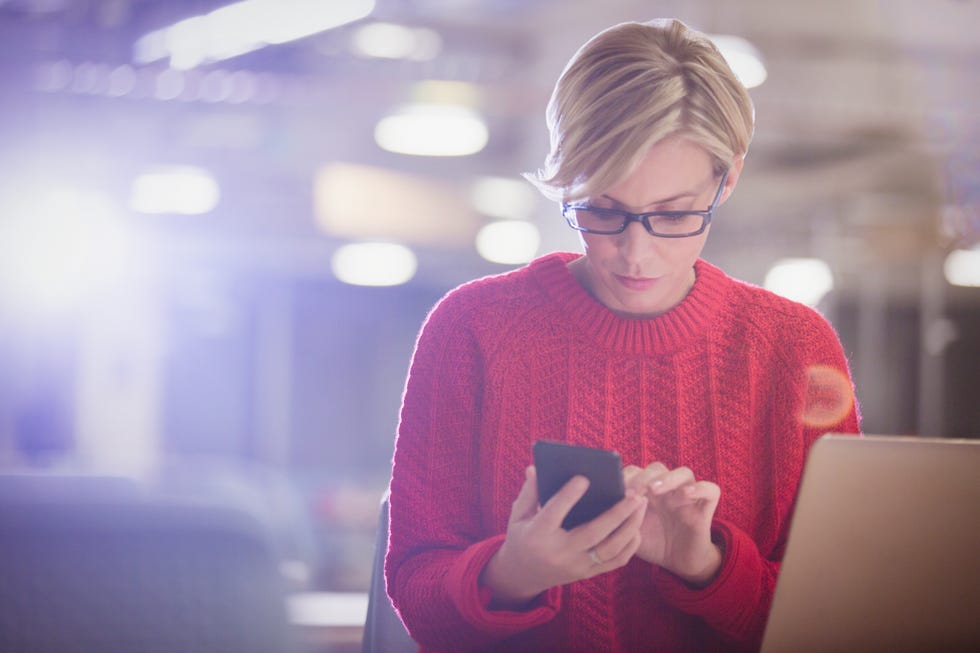 Businesswoman working late at laptop, texting with cell phone in dark office